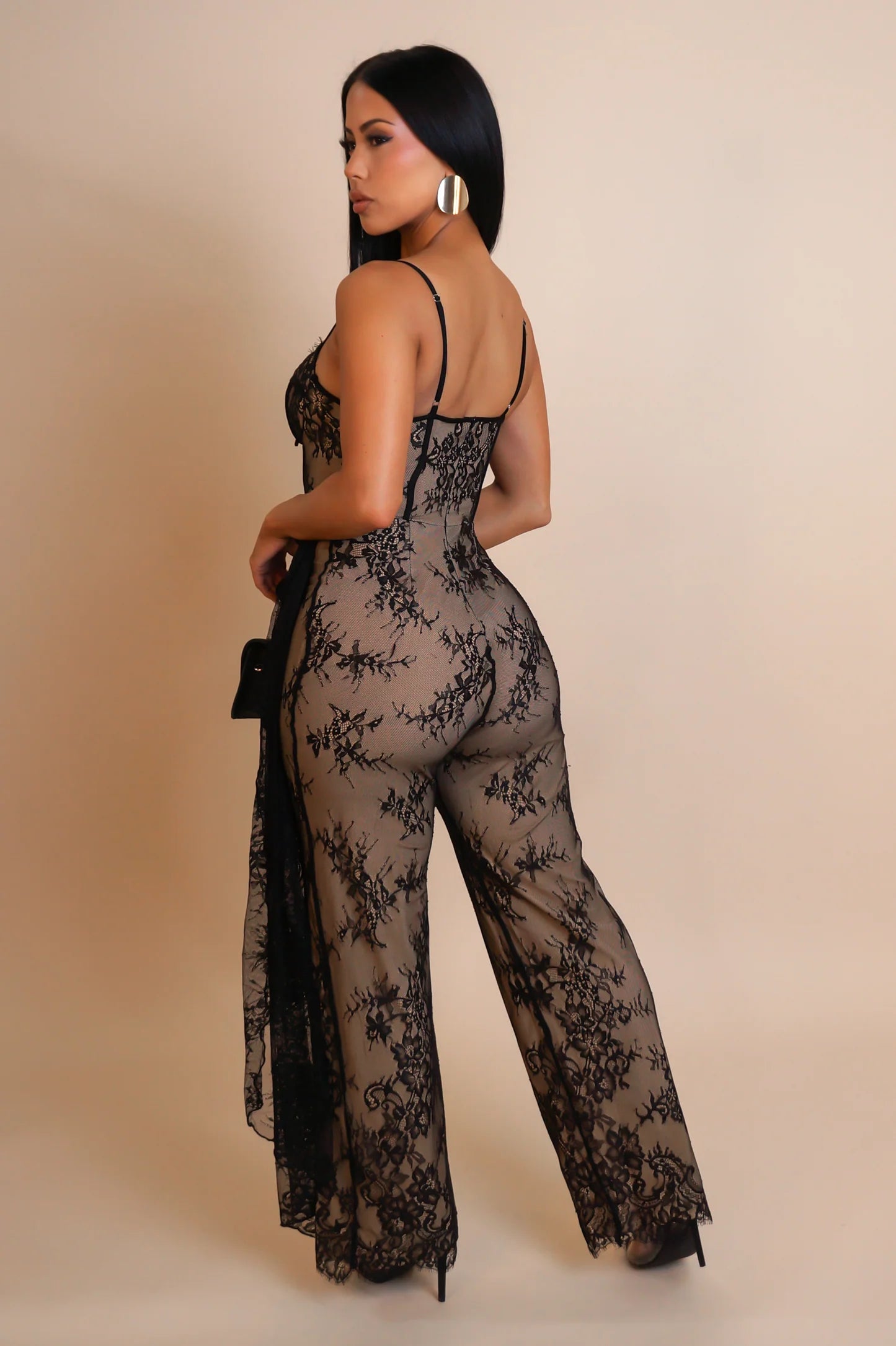 A model wearing a black sheer lace jumpsuit with floral detailing, featuring a corset-style bustier top with underwire cups and adjustable straps, high-waisted fit with a wide-leg silhouette, and a hidden back zipper.